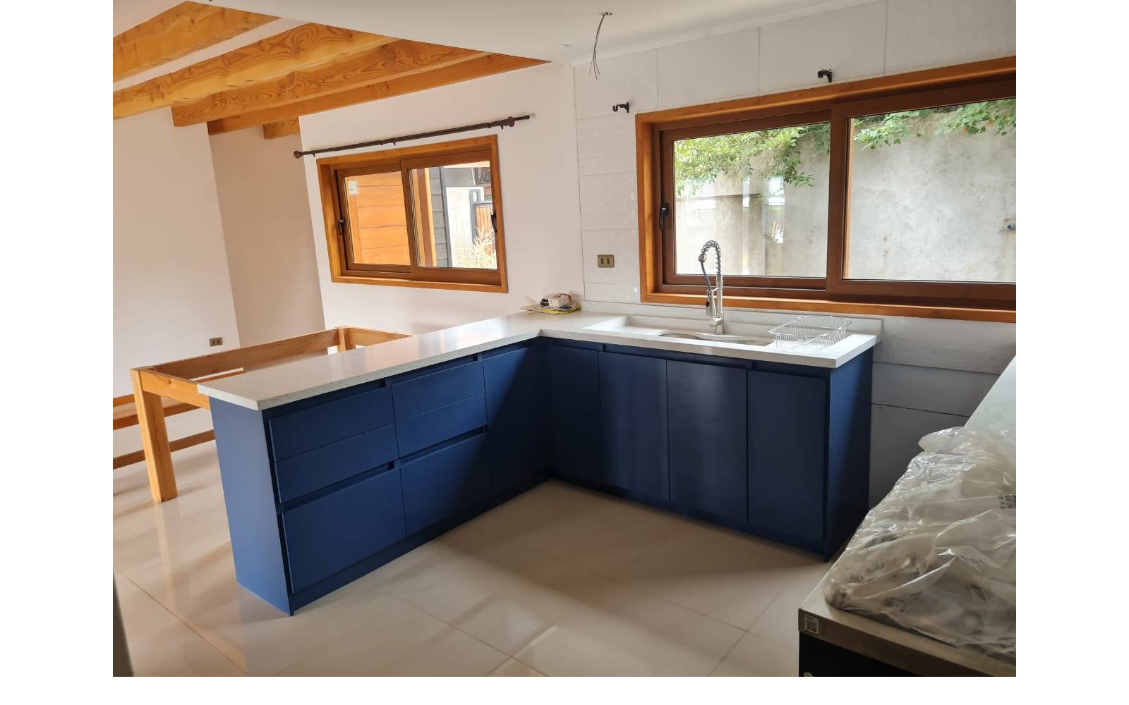 Cocina con muebles azules, cubierta blanca y ventanas de madera.