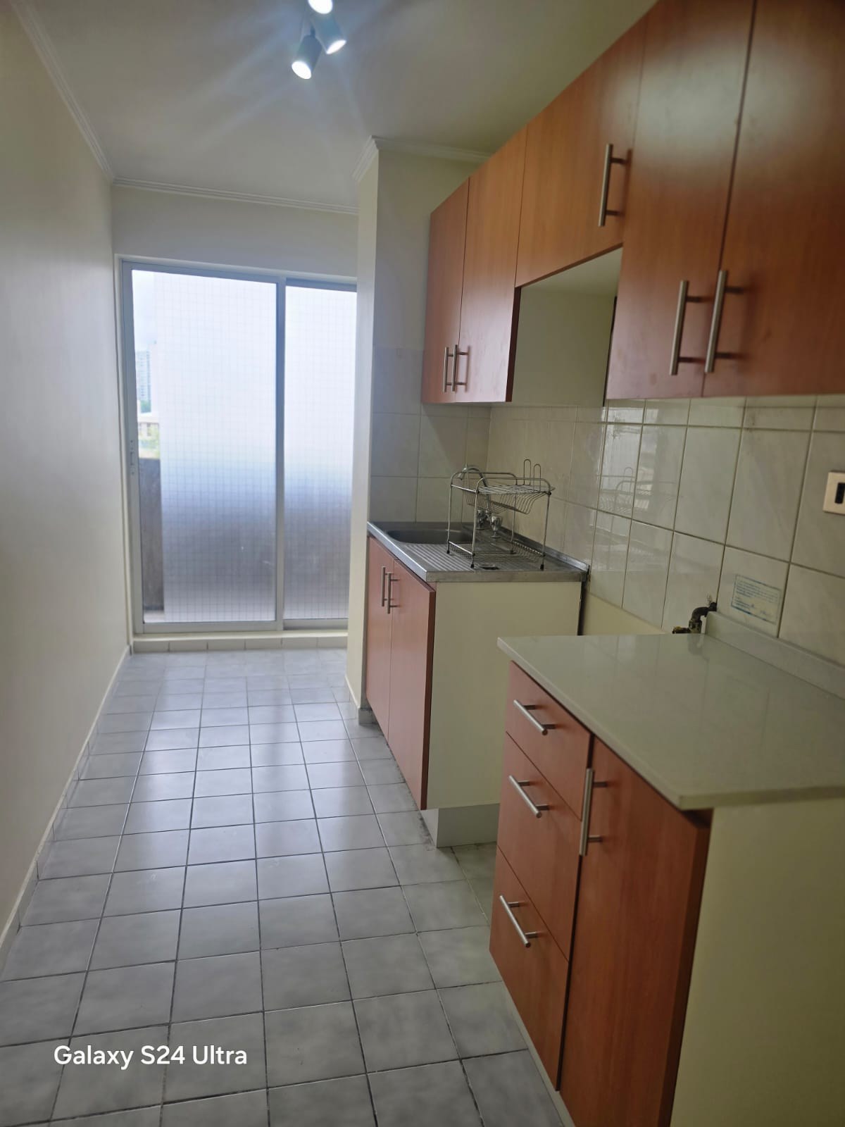 Cocina alargada con muebles de madera, lavaplatos y logia al fondo